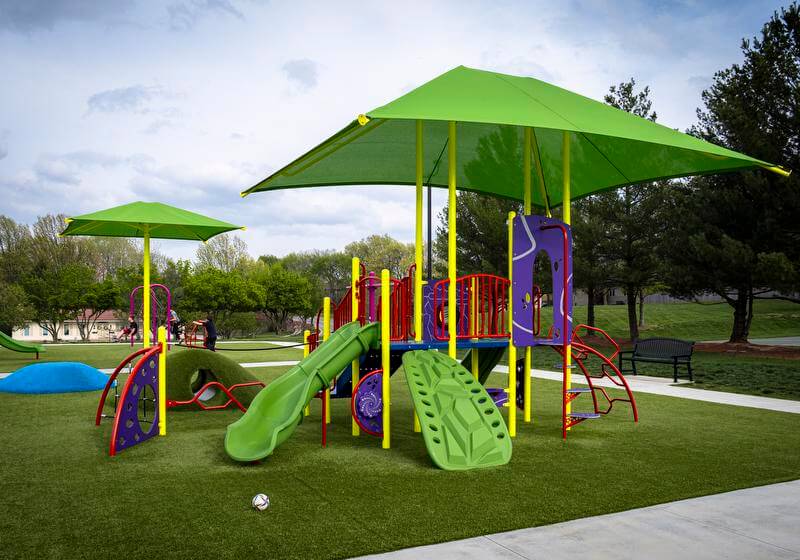 Vibrant playground equipment with slides and climbers on green turf, shaded by oversized umbrellas.