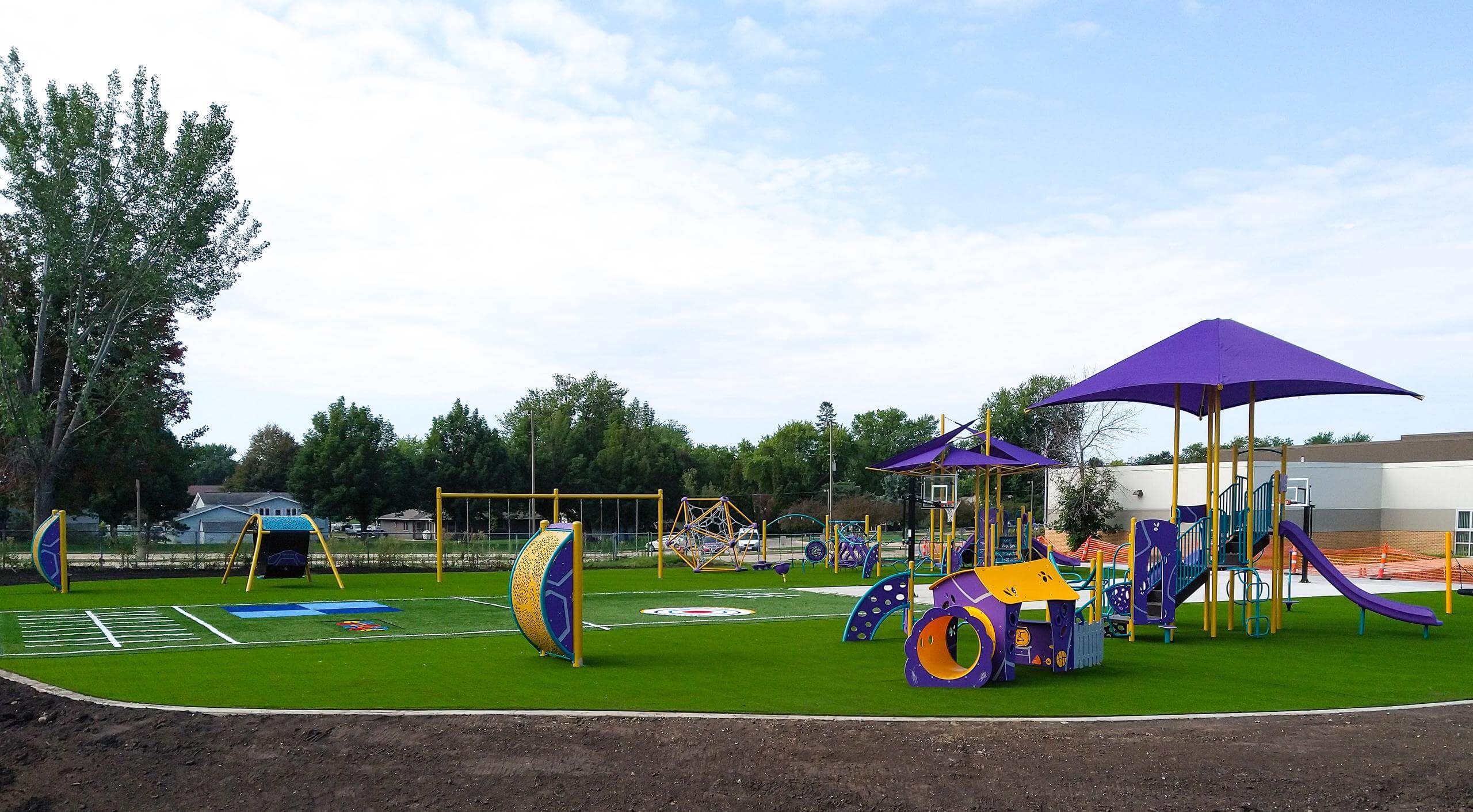 Spacious playground with colorful play structures, shaded areas, and synthetic grass surface.