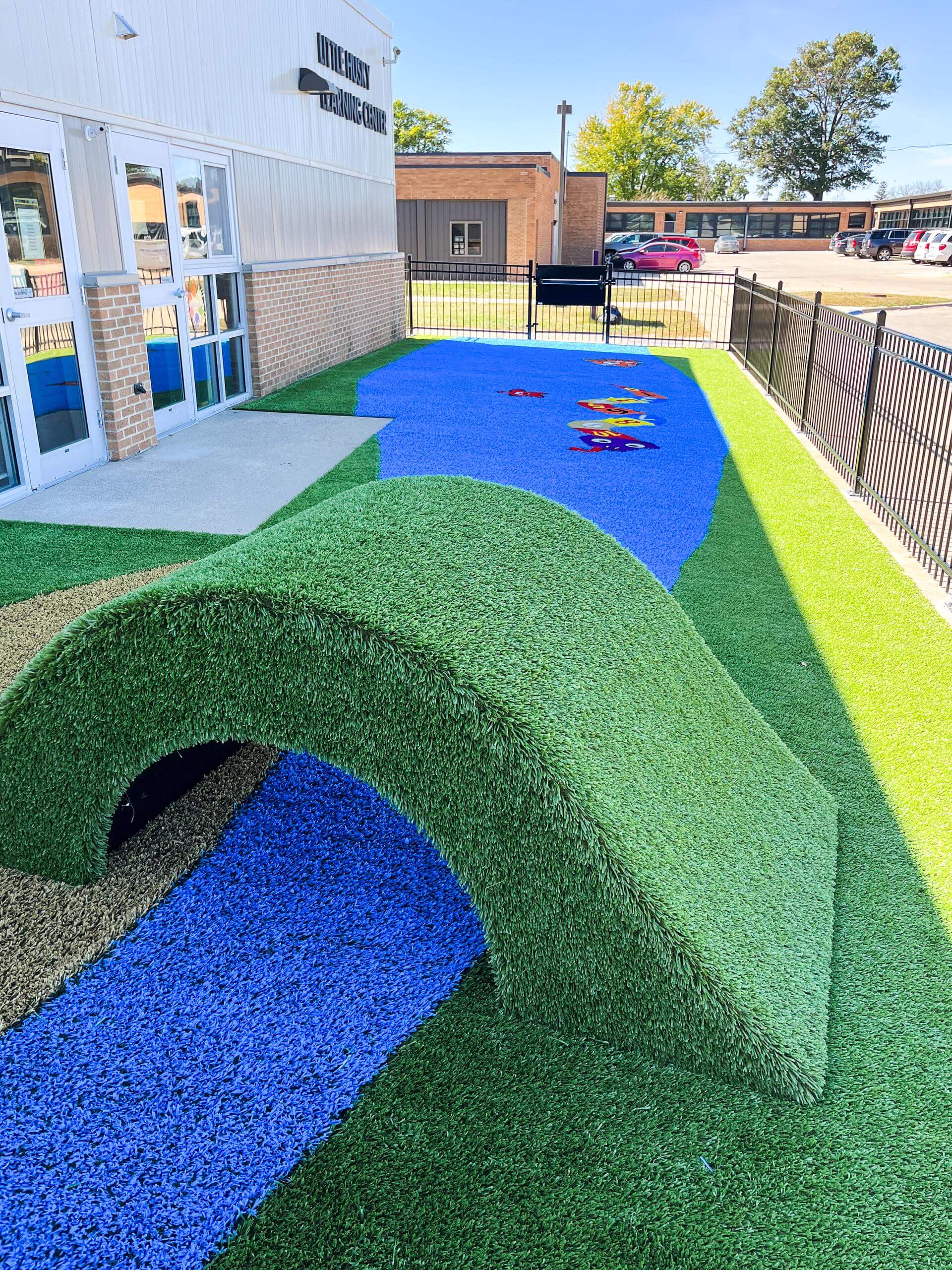 Colorful tunnel entrance on a playground with blue synthetic stream and green turf hills.