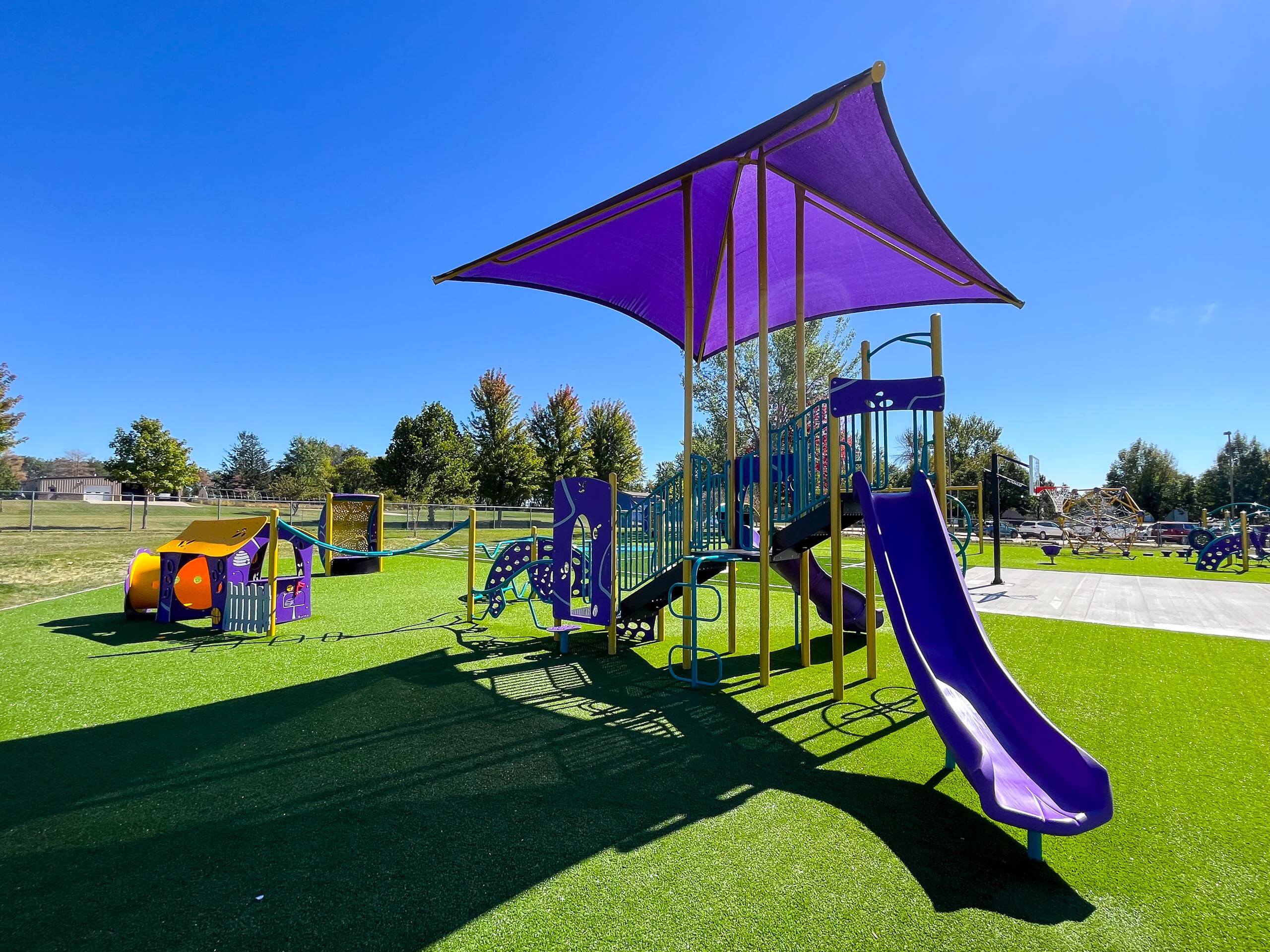 Sunny playground with vibrant purple shades, slides, and climbing structures on green artificial grass.