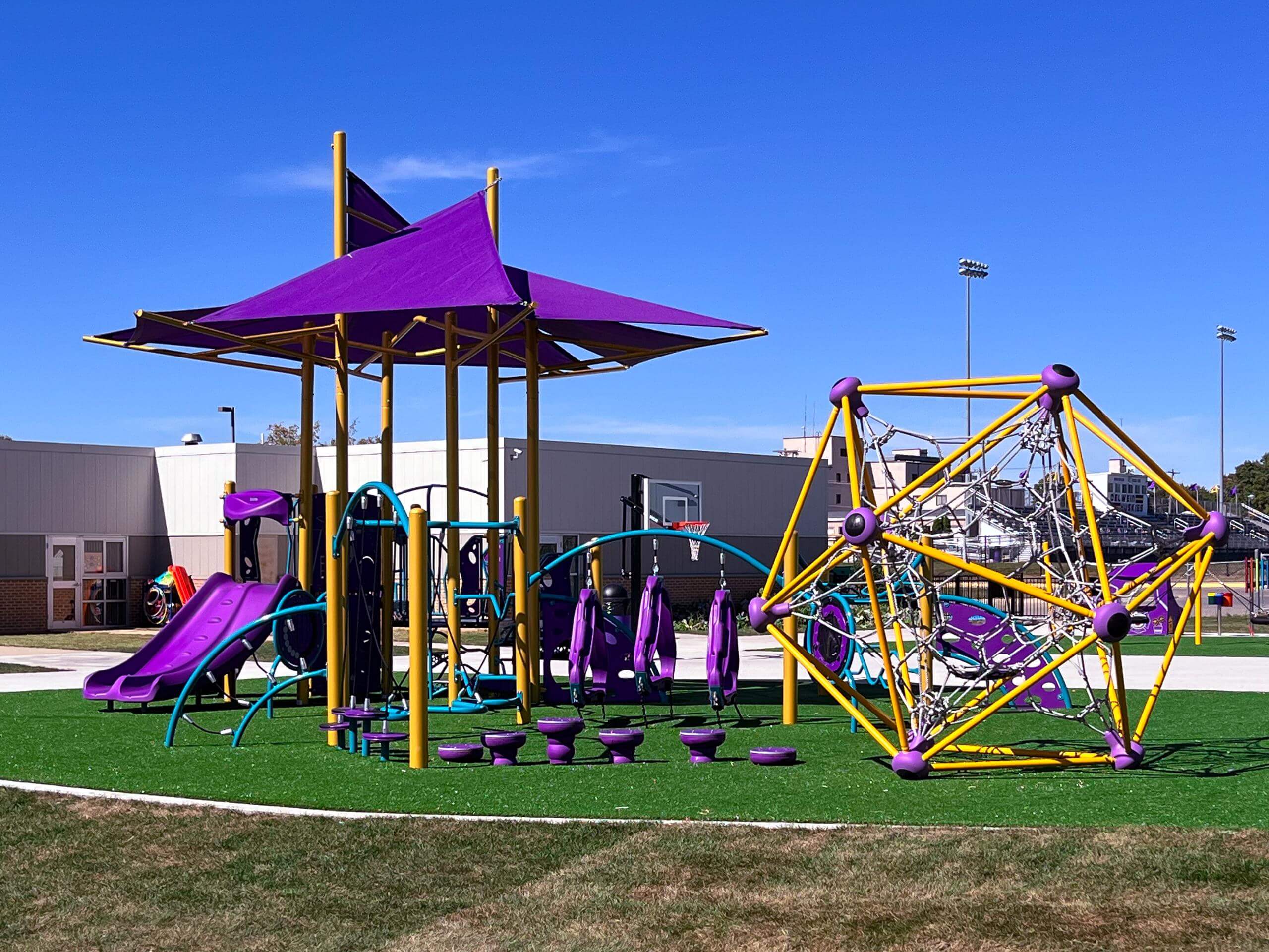 Play area with yellow climbing dome, purple slides, and shaded platforms on synthetic turf.