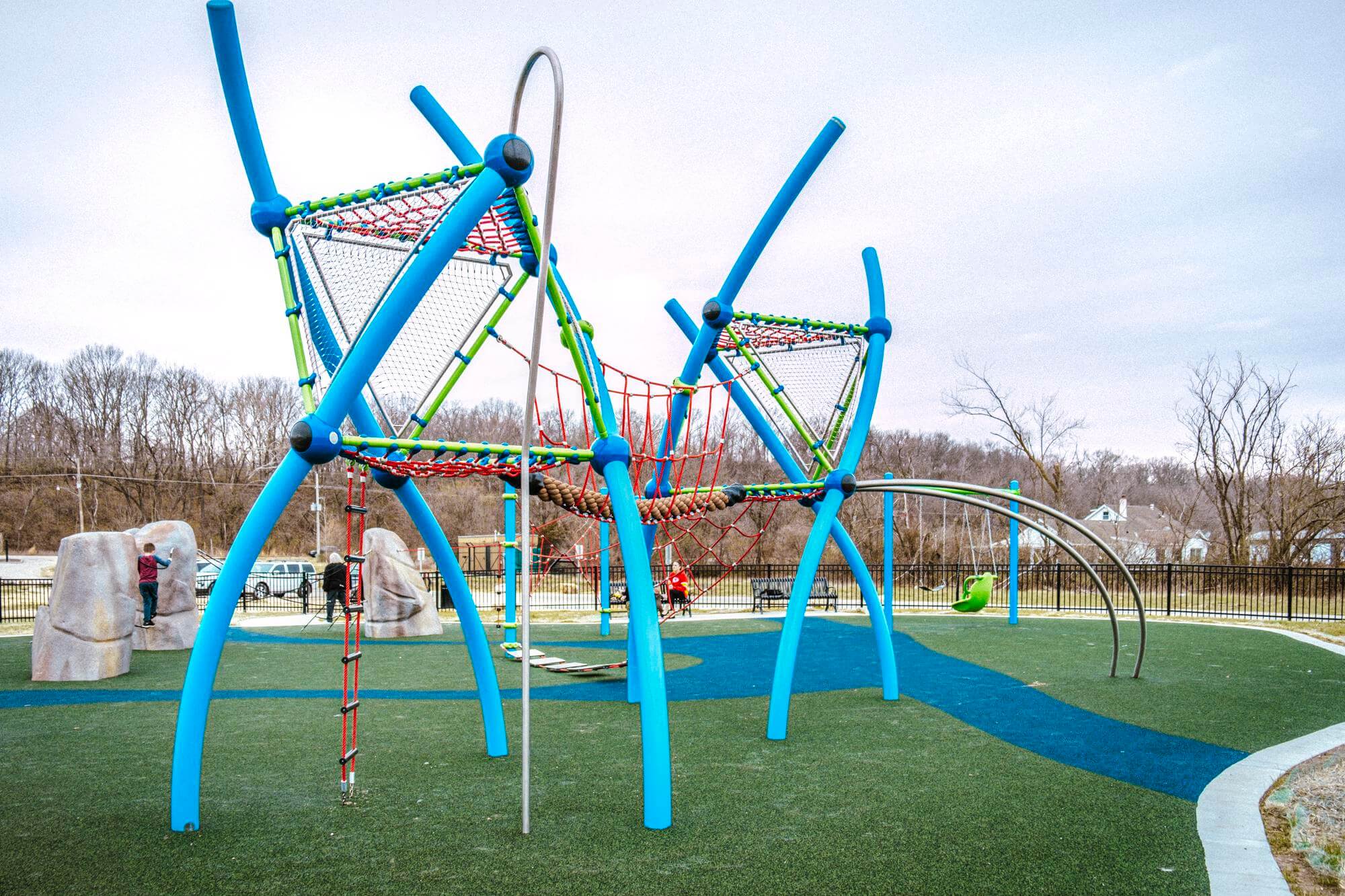 playground featuring a mix of green synthetic turf and blue pathways. In the foreground, three sculptural boulders equipped with climbing ropes and ladders are prominently displayed.