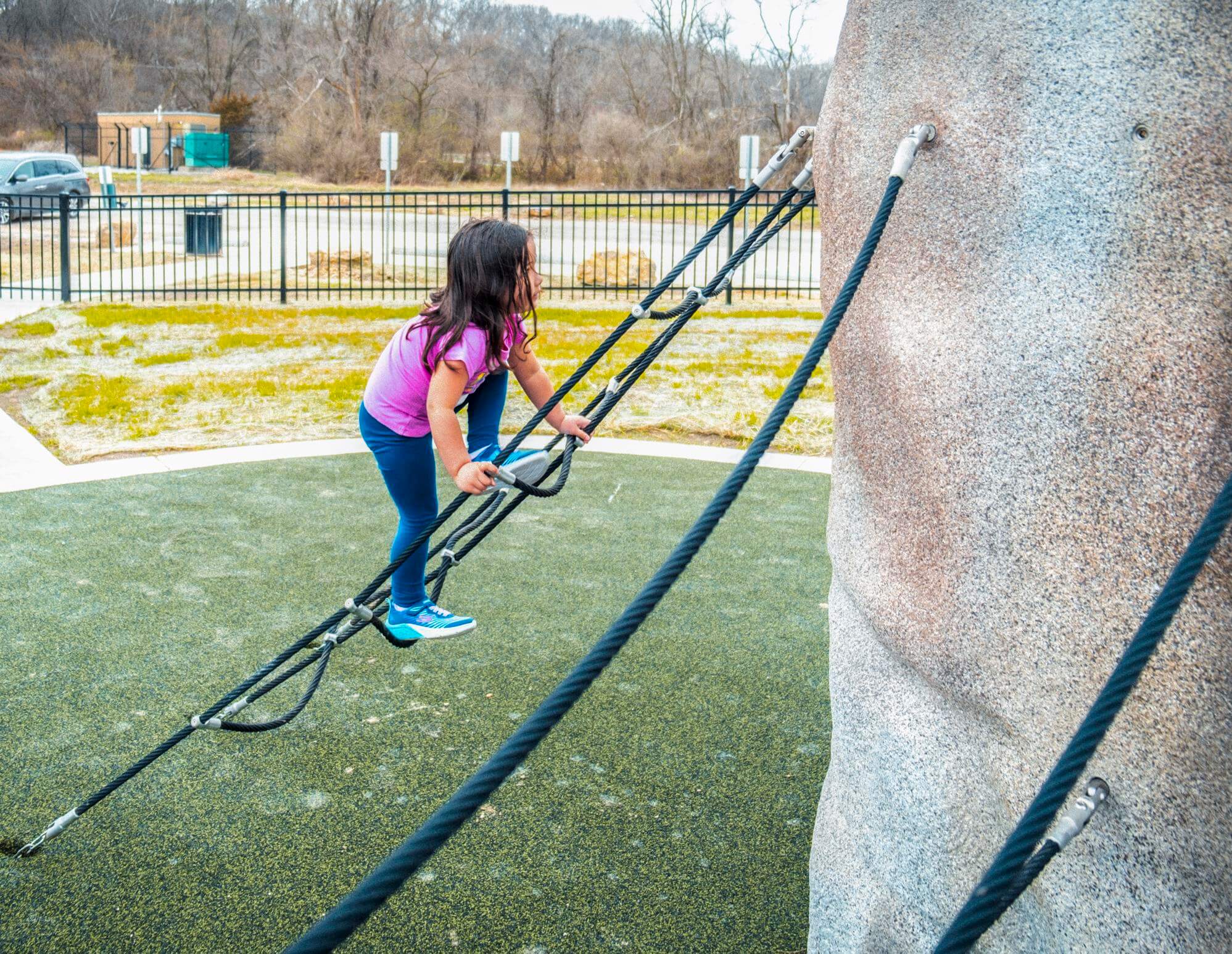 A child is focused on climbing a rope structure connected to a large, lifelike boulder in a playground. The synthetic green turf around her provides a safe playing surface.