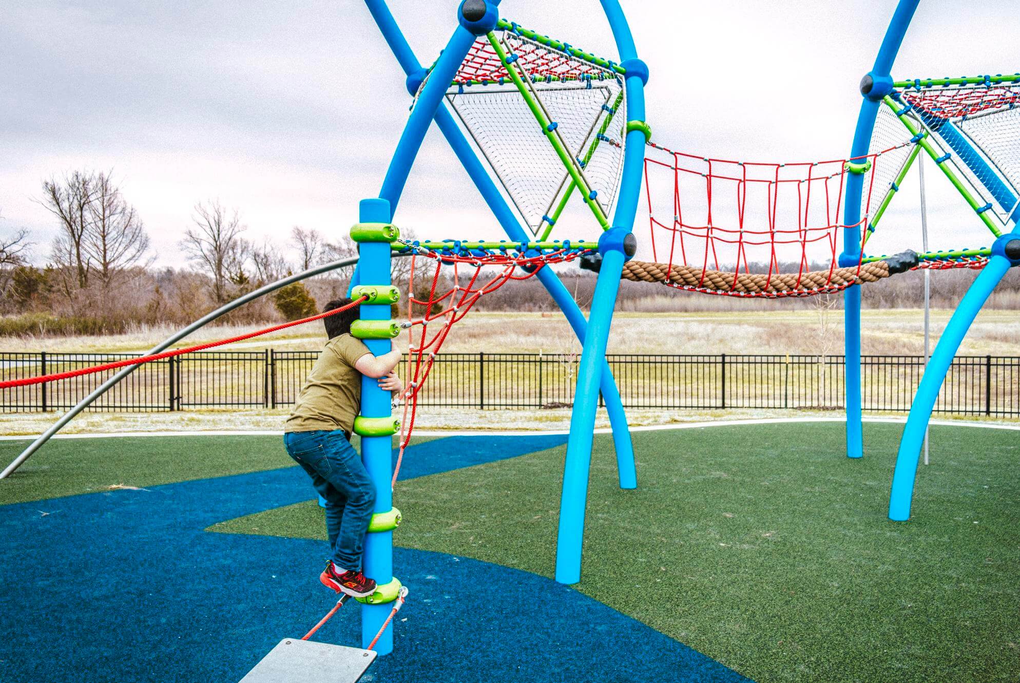 A child in a green shirt climbs a blue climbing frame with rope elements, on a playground with a mixture of blue and green synthetic grass.