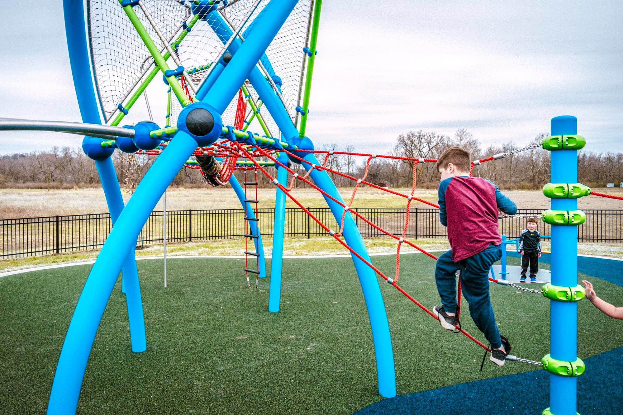 A child climbs a red rope on a blue climbing frame, with another child observing from below, in a playground with diverse play structures.