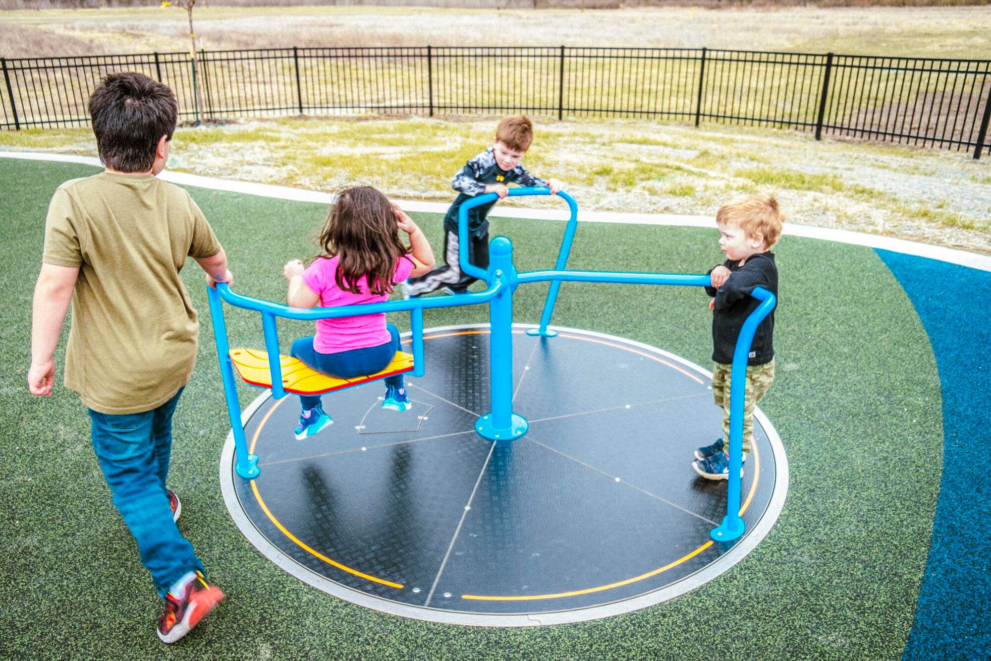 Children play on a round, spinning platform with blue handrails on a playground with green synthetic turf and a blue path.