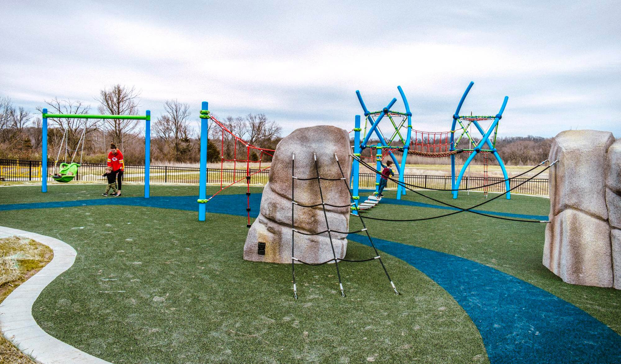A playground featuring a series of sculptural climbing boulders interconnected with rope bridges, against a backdrop of a vibrant rope climbing structure.