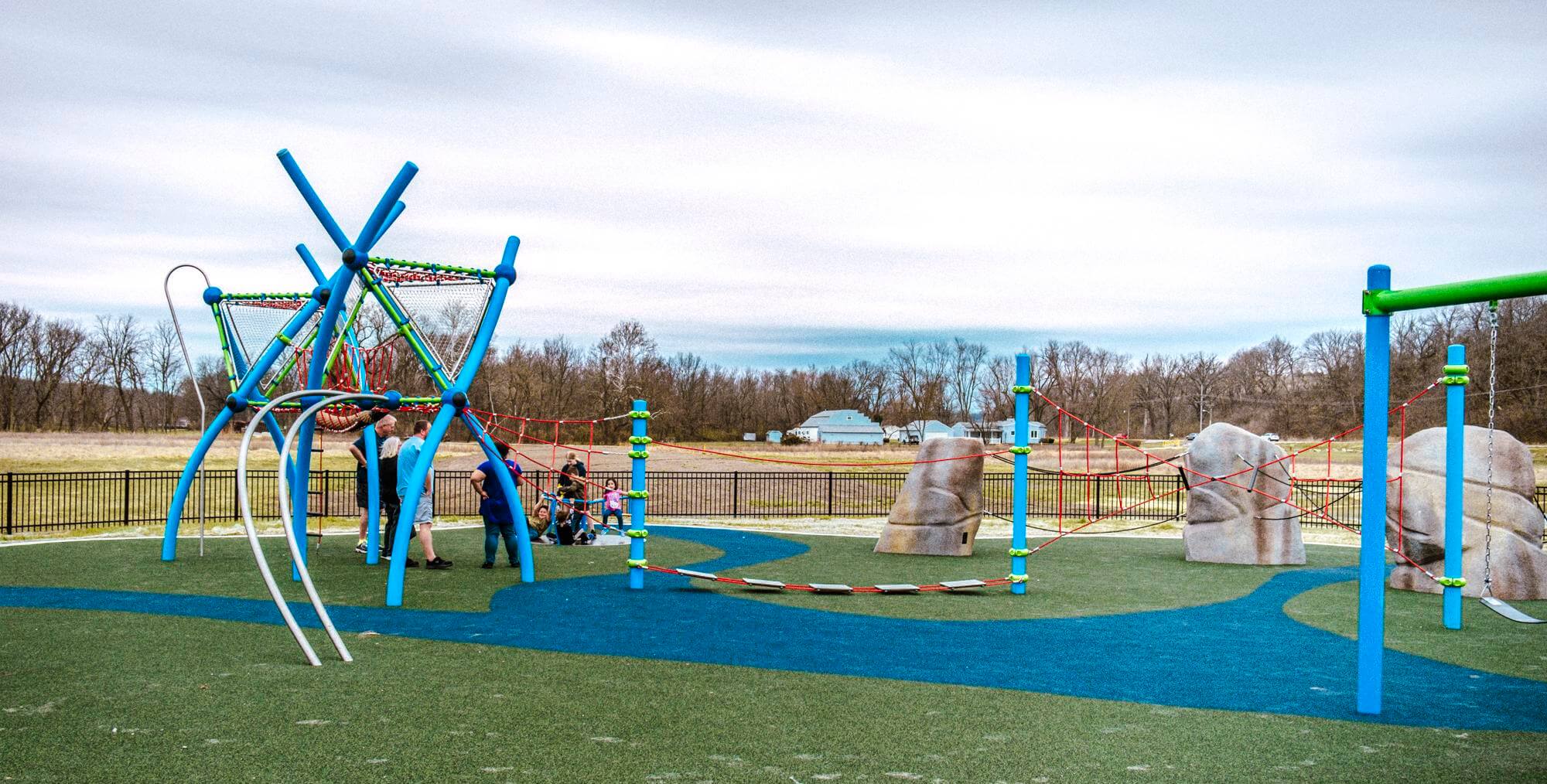 A playground landscape showing a rope bridge connecting two climbing boulders with a complex climbing structure in the background.
