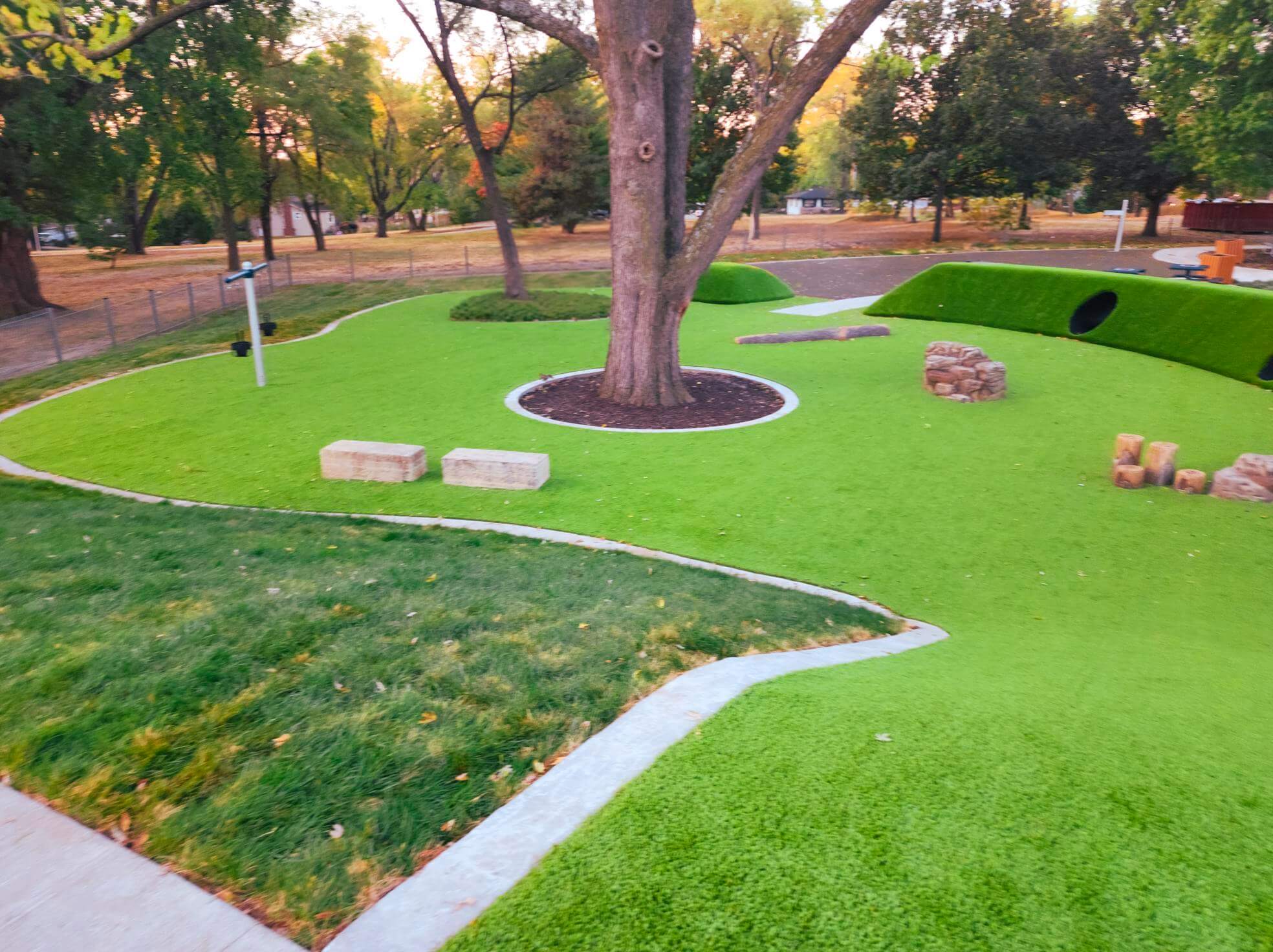 A sprawling playground featuring undulating green artificial hills, and a central tree.