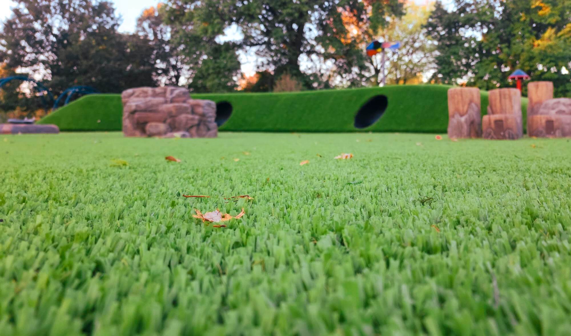 A playground with artificial green turf and scattered leaves, featuring play structures such as tunnels in artificial hills, captured in soft lighting.
