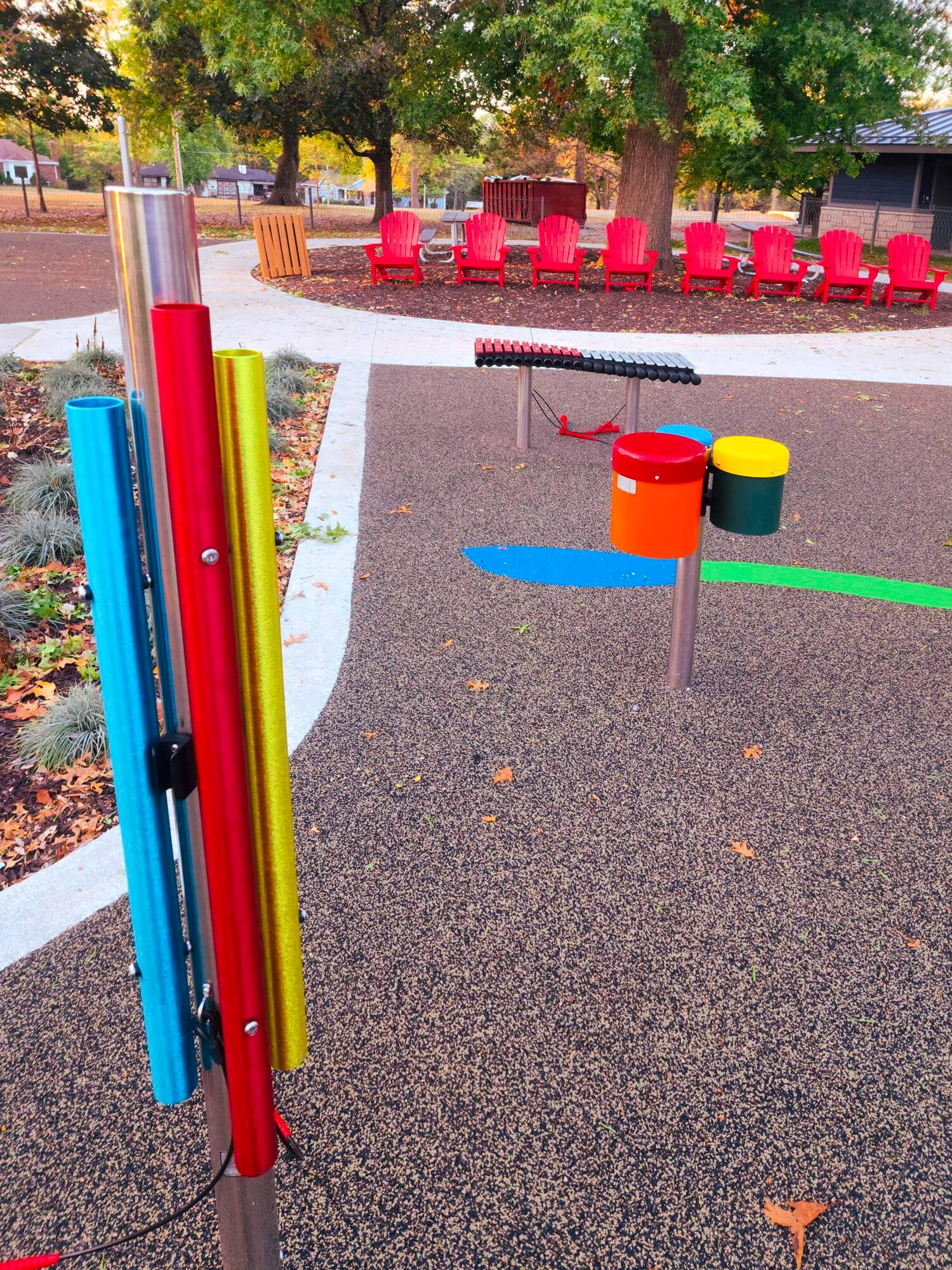 A vibrant playground with colorful chimes and drums in the foreground, and red Adirondack chairs arranged around a fire pit in the background, set against an autumnal tree line.