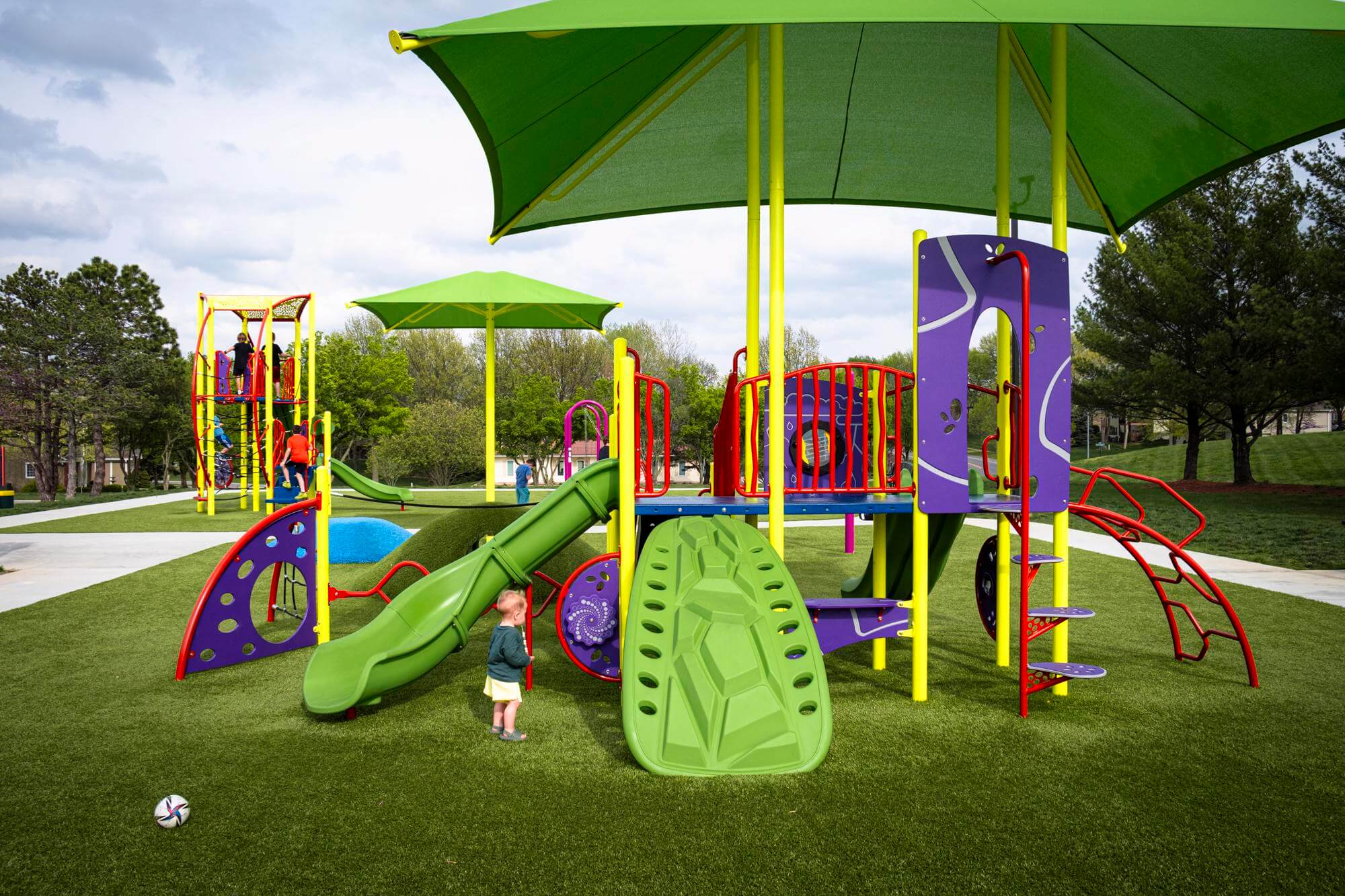 Colorful playground with green synthetic turf, featuring a slide and sunshades, with a child standing nearby.