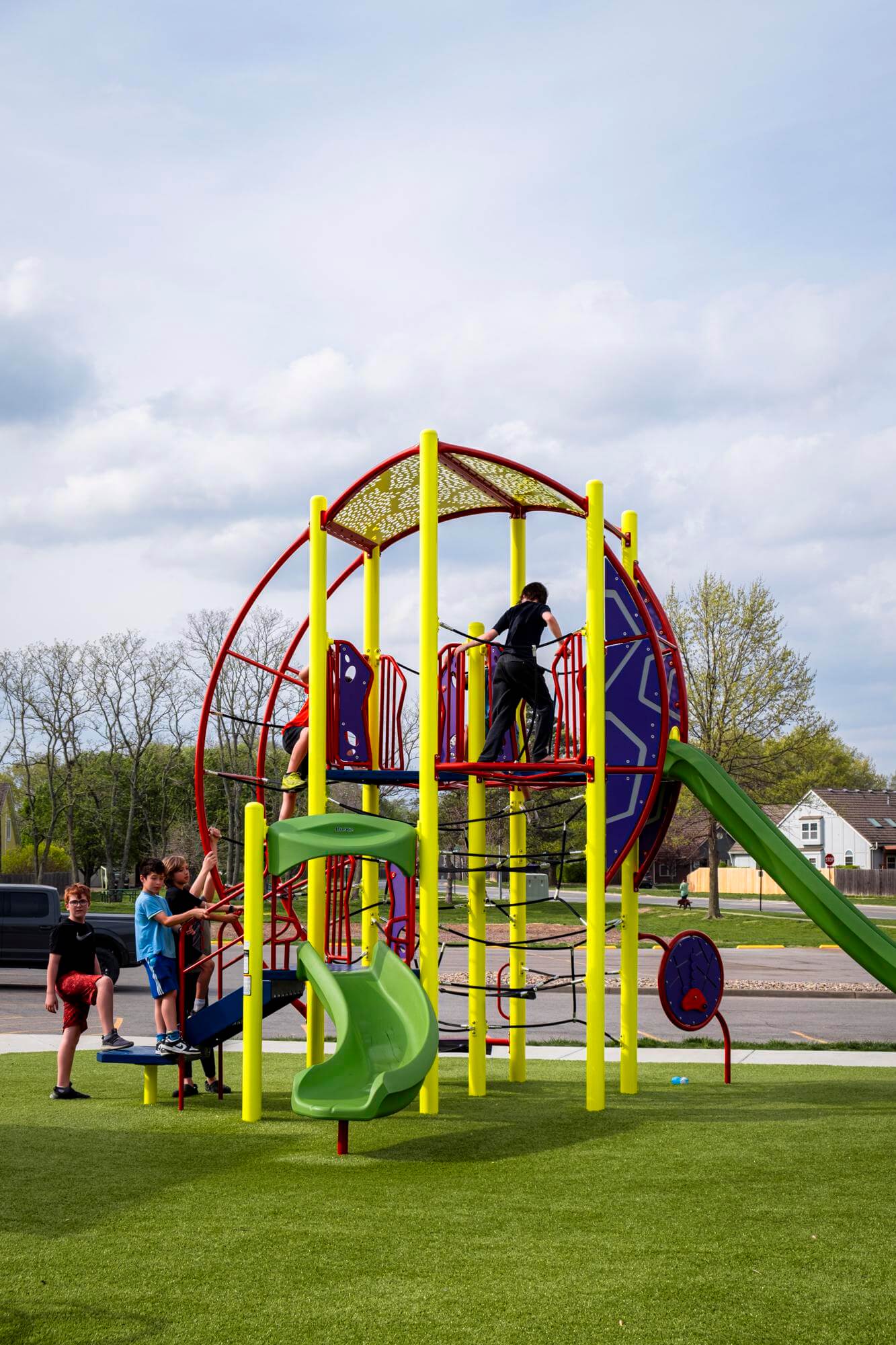 Children playing on a playground with a climbing tower and slide on a lush green artificial turf.
