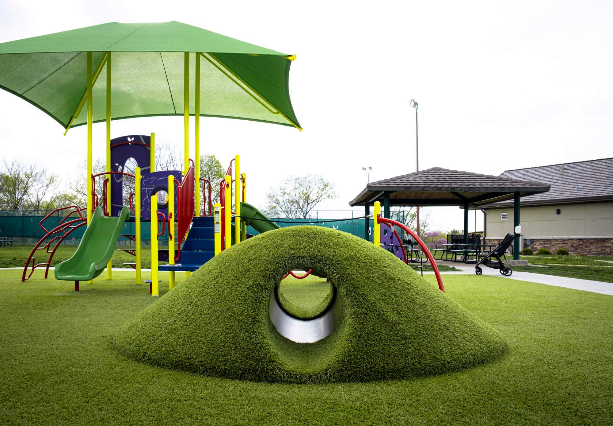 Colorful playground with vibrant play structures, oversized umbrella for shade, and tunnel turf mound.