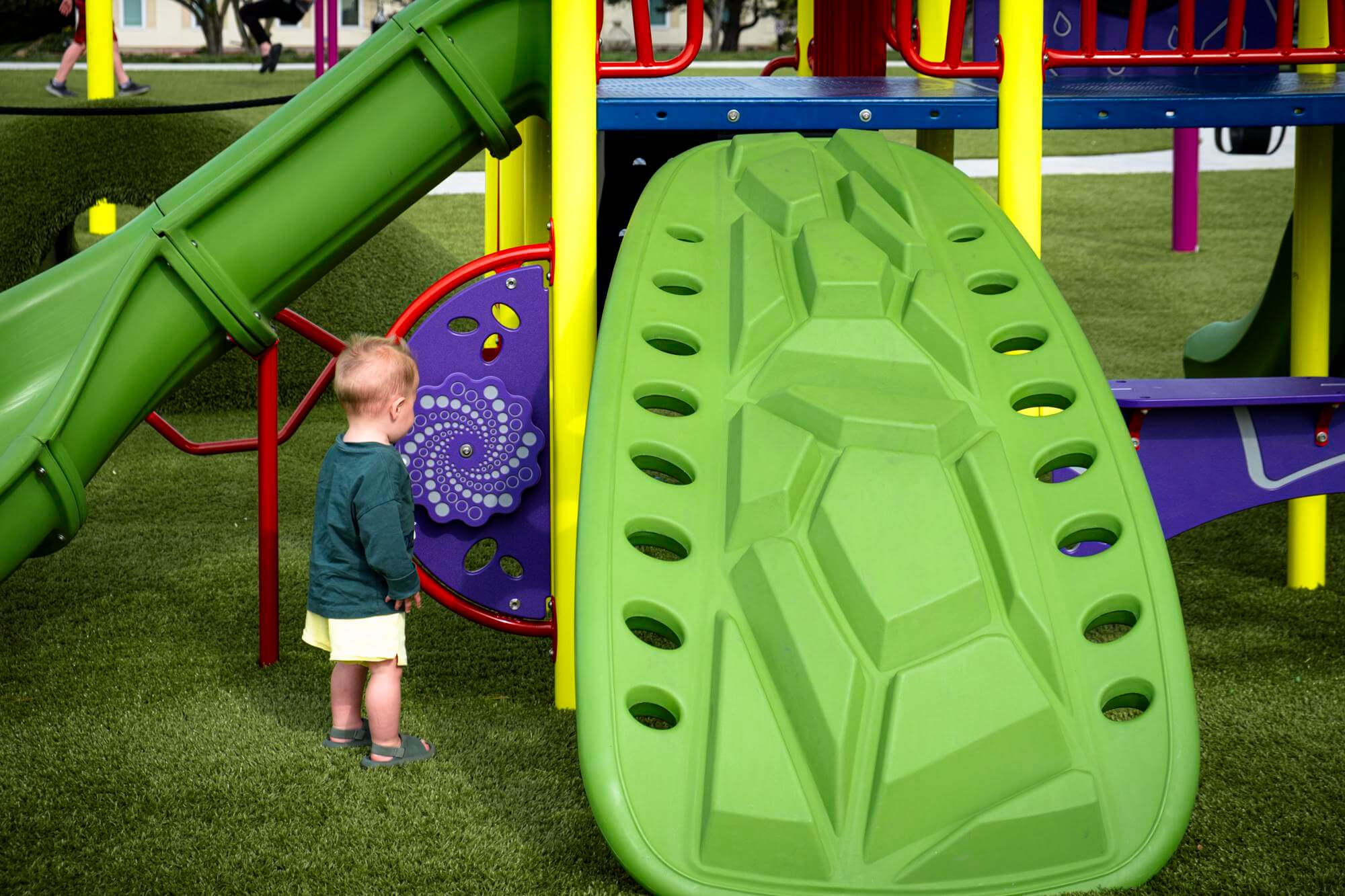 Child looking at an interactive play panel on a playground with artificial grass and a climbing structure.