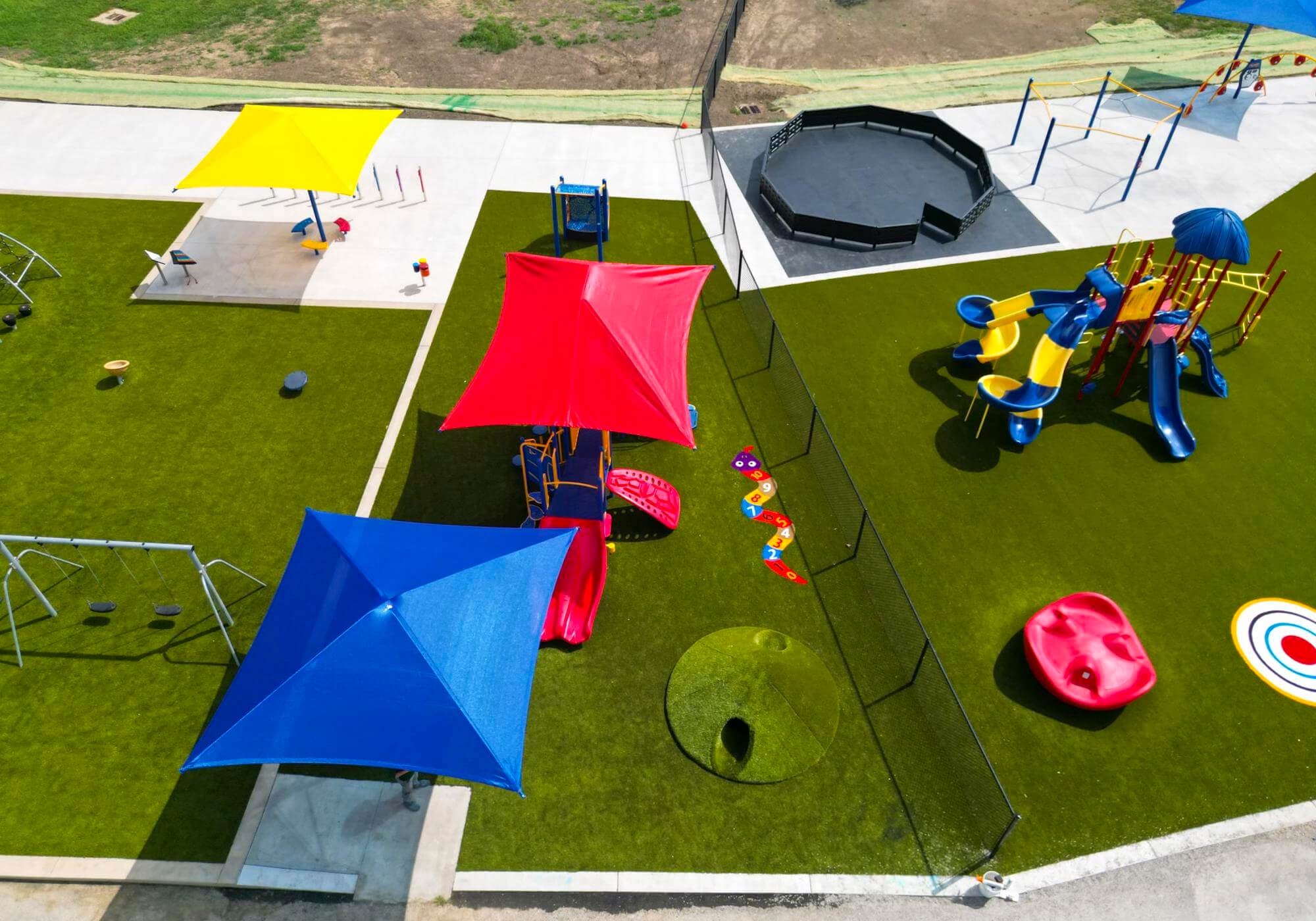 An aerial view of a playground with various play structures, and bright sunshades, surrounded by a green field.