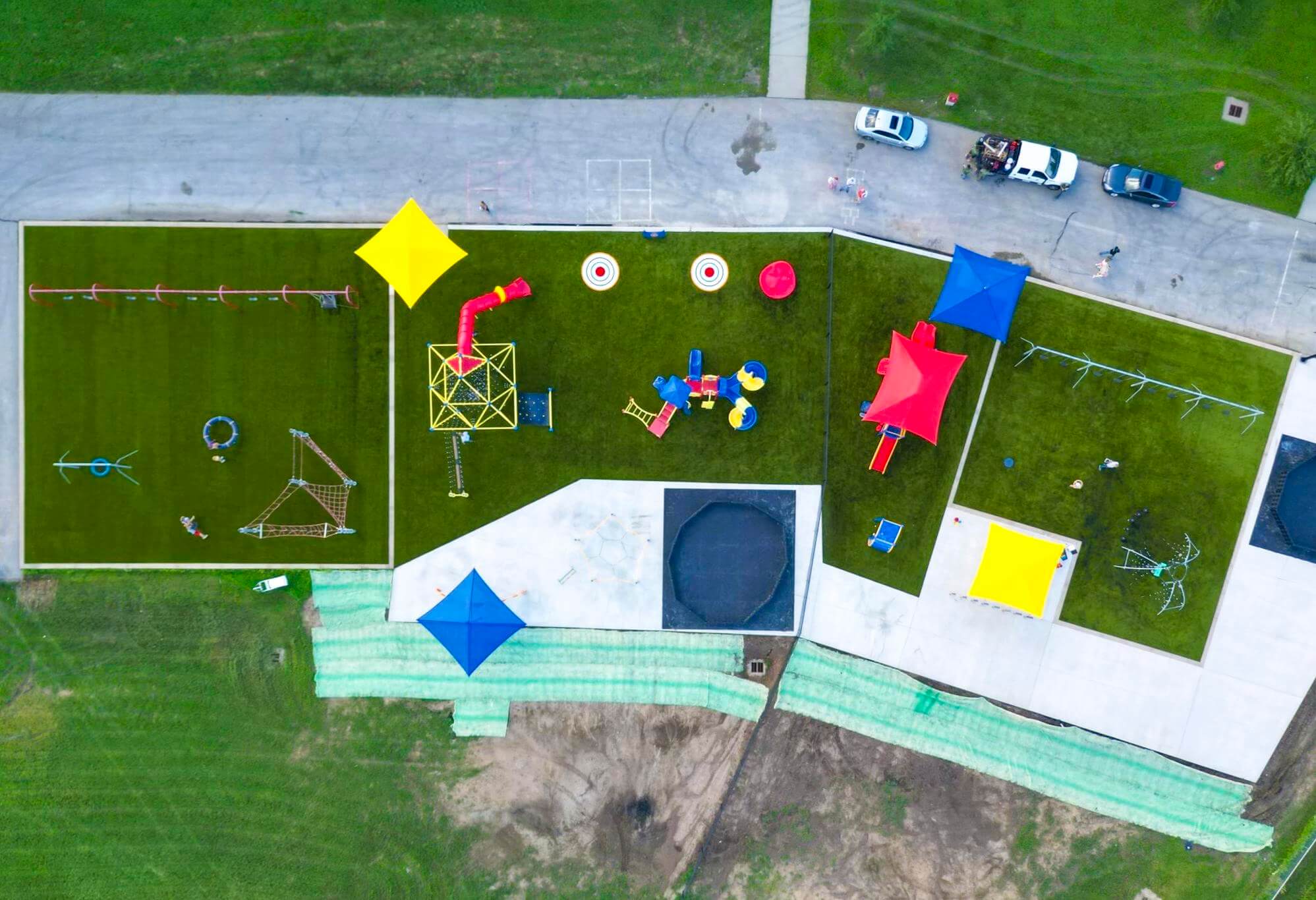 Top-down aerial shot of a playground with geometric sunshades, a sports court, and play equipment on a sunny day.
