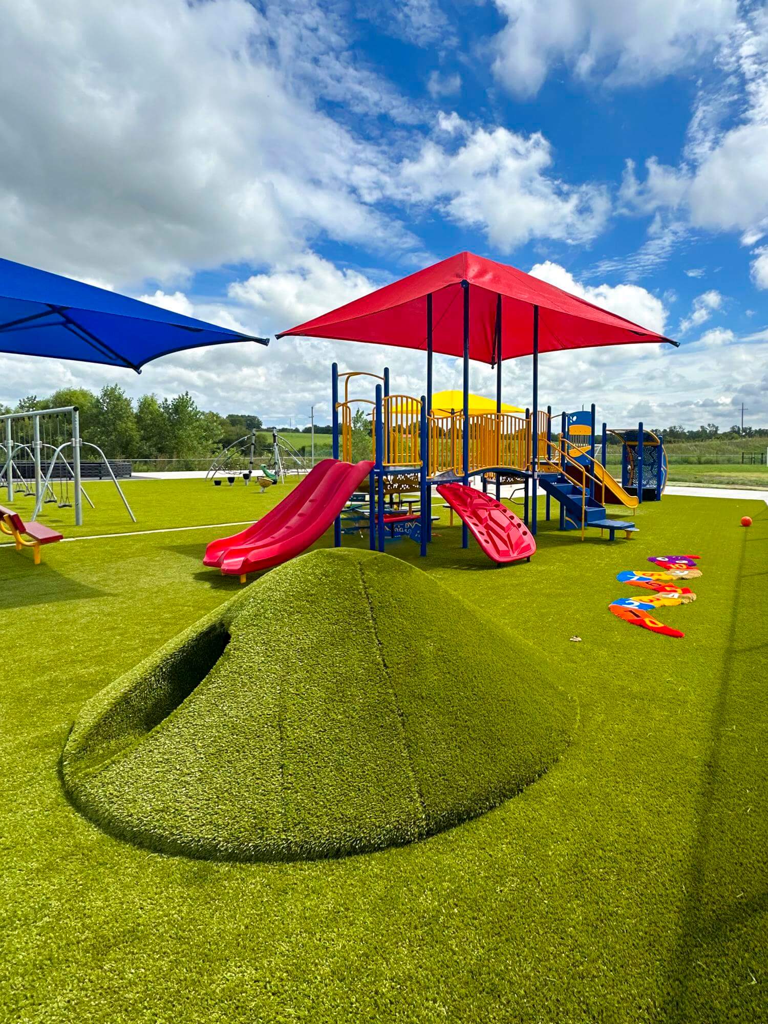 Close-up of a playground with artificial turf mounds, red and blue sunshades, and a variety of play equipment.
