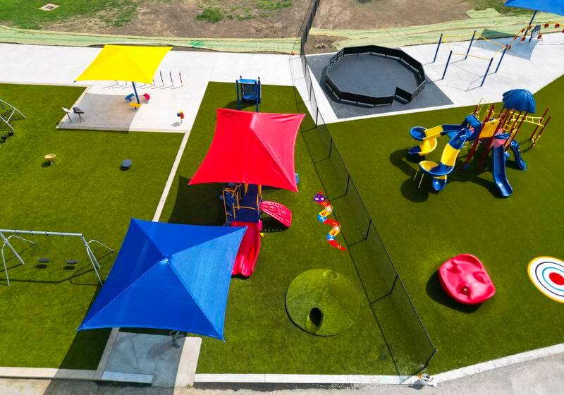 An aerial view of a playground with various play structures, and bright sunshades, surrounded by a green field.